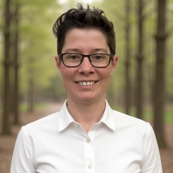 Person mit Brille, lächelnd, steht in einem Wald mit grünen Bäumen im Hintergrund.