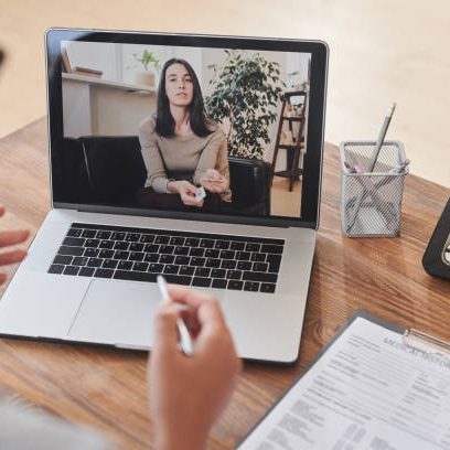 Person spricht in einem Videoanruf auf einem Laptop, Notizen und Stift liegen daneben.